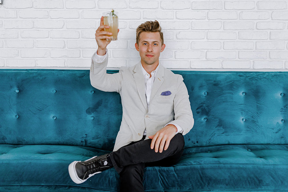 Man in suit raises drinking glass to salute while sitting on blue couch.