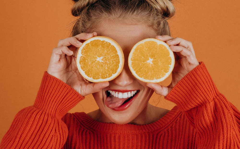 Girl in red sweater holding orange halves over her eyes on orange background.